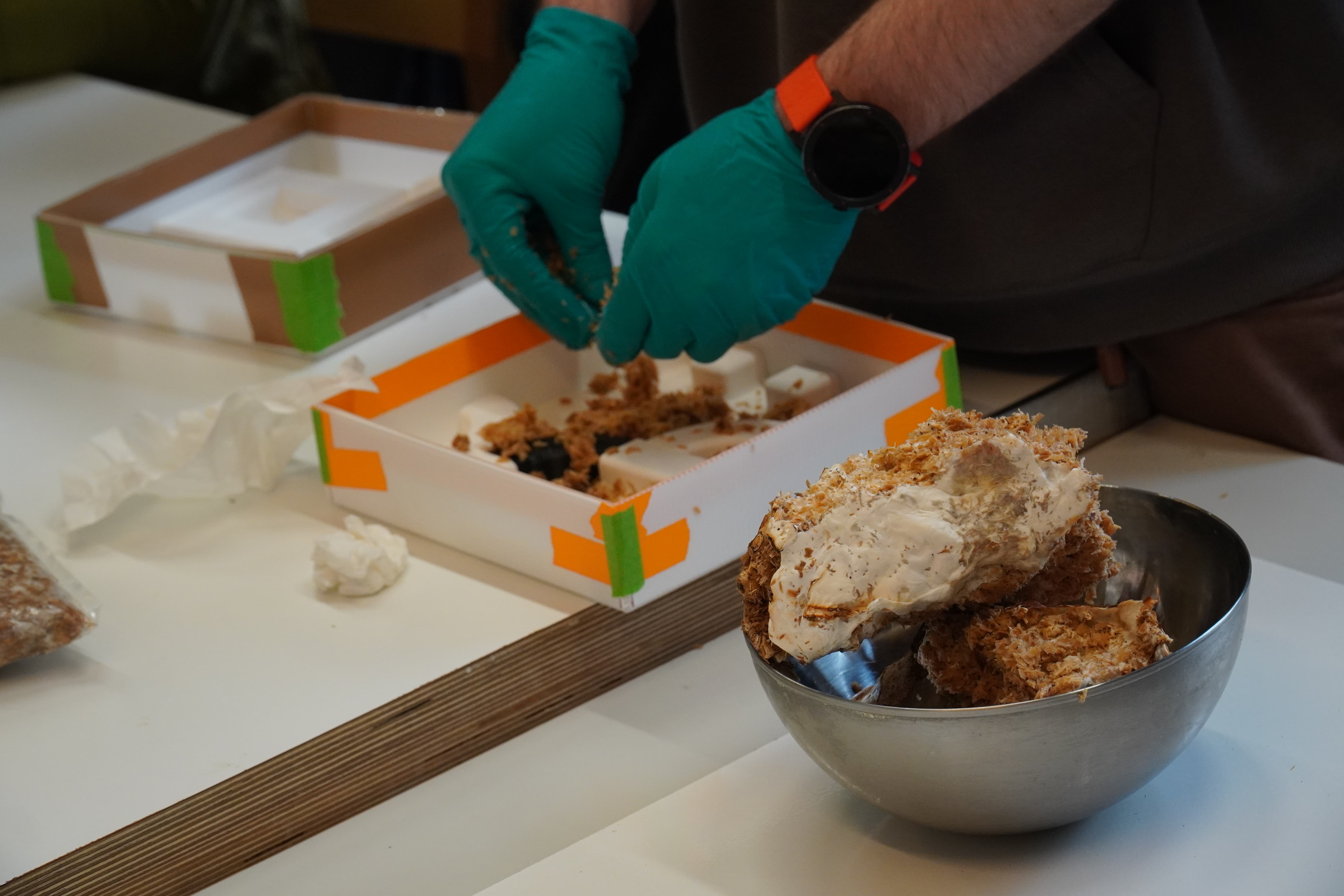 A gloved hand is taking apart and fitting in the mycelium chunks (fed with woodchips) into their mould.